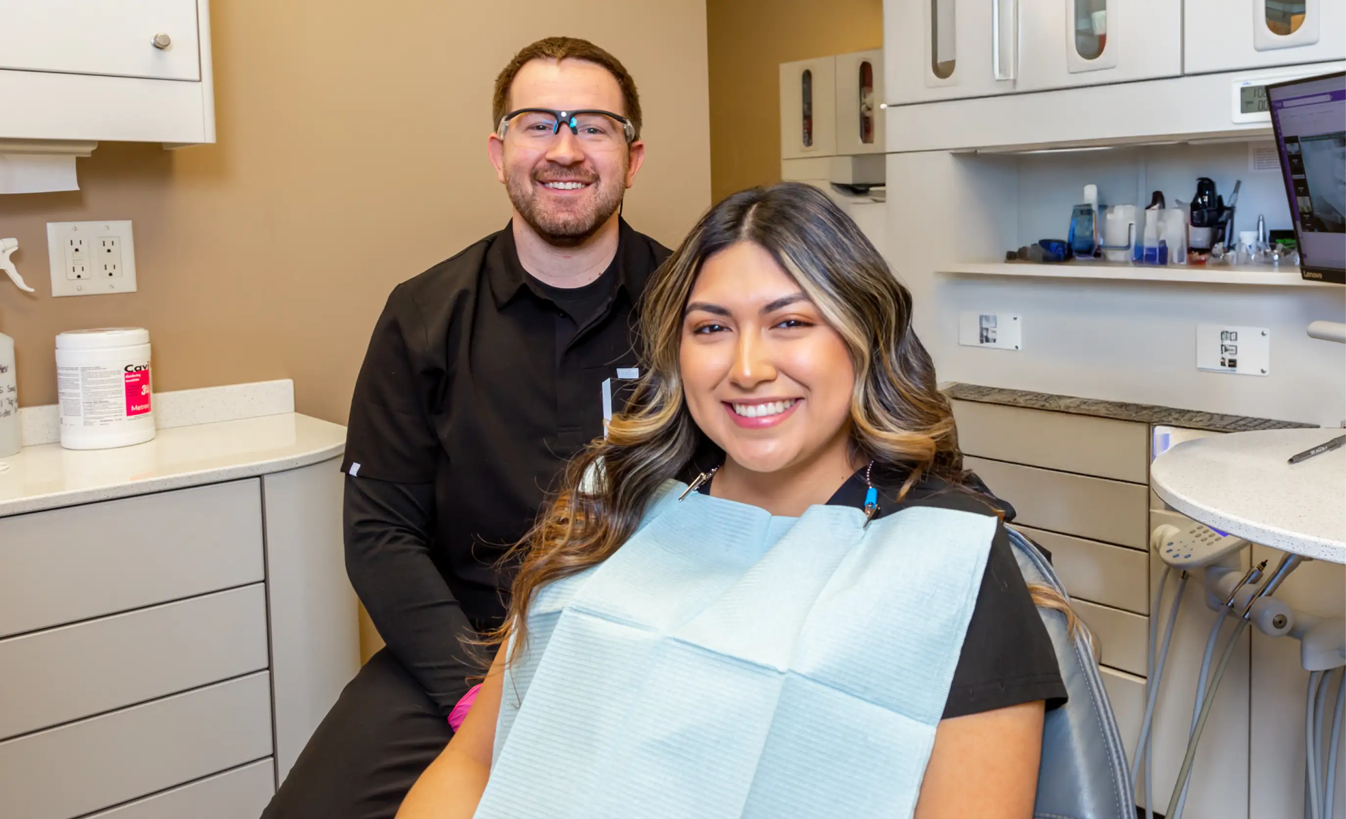 Smiling female dental patient seated in chair with male dentist wearing protective glasses standing behind her in a dental office.