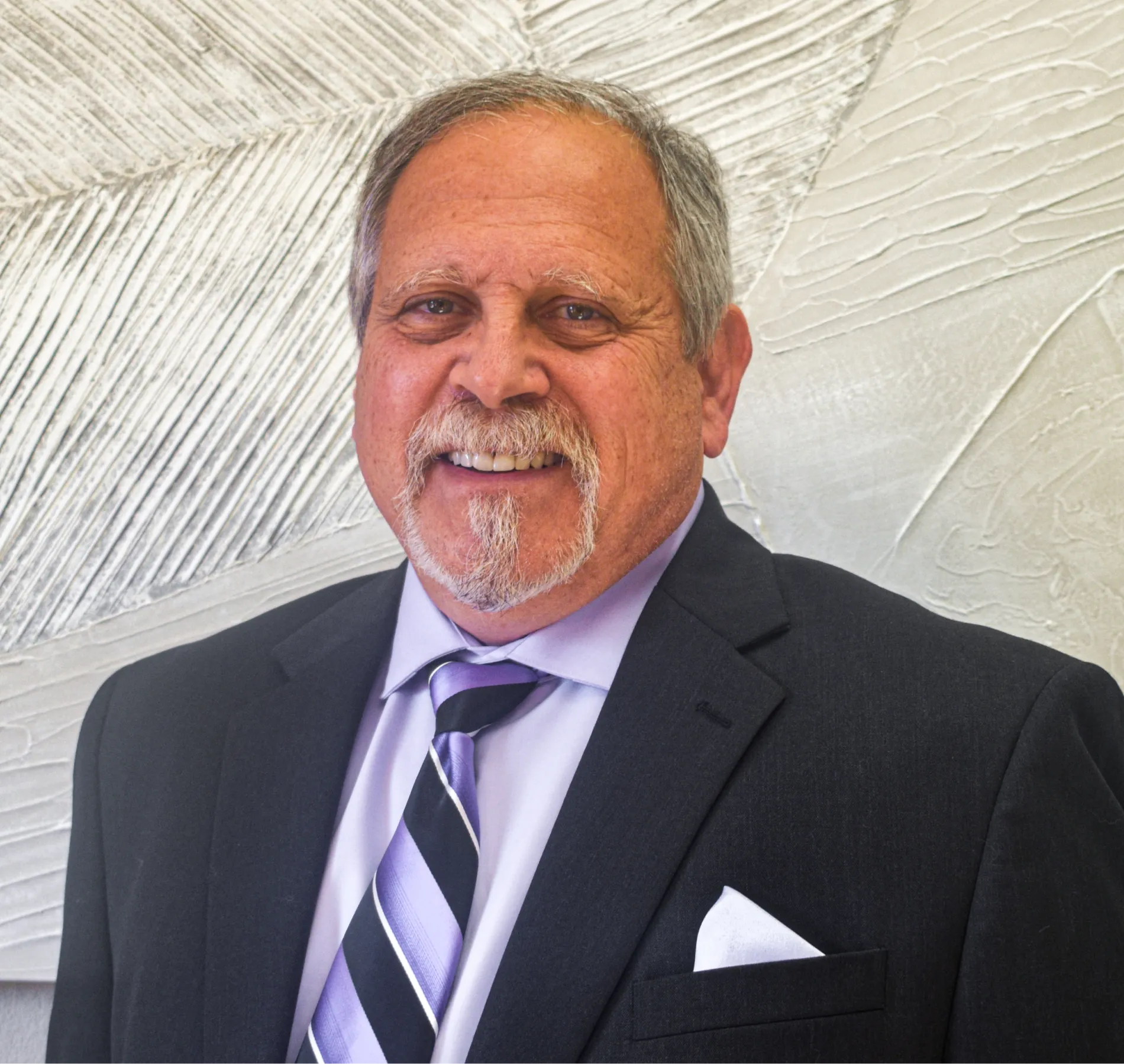 Smiling older man with gray hair and goatee wearing a dark suit, light purple shirt, and striped purple tie.