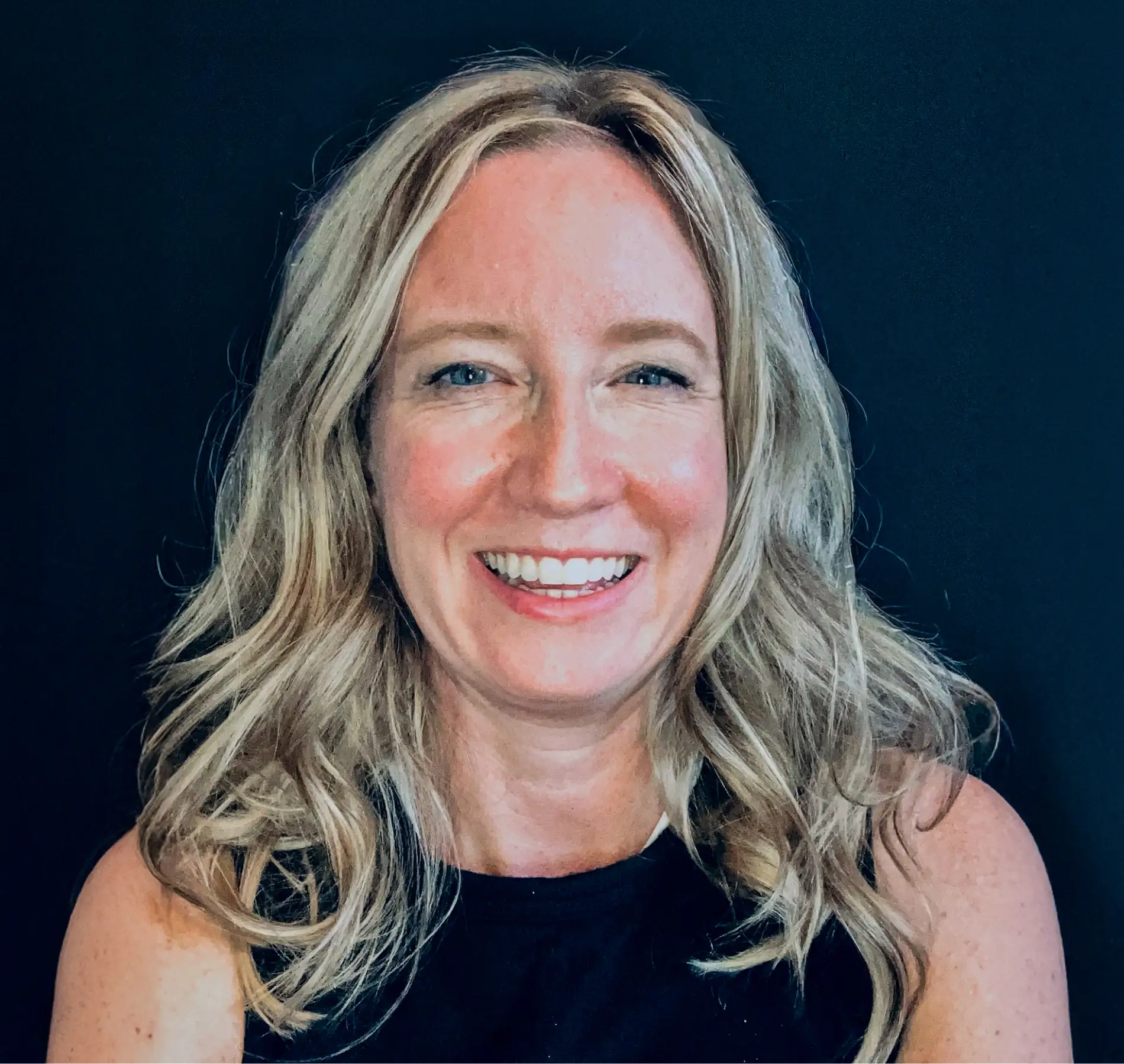 Smiling woman with wavy blonde hair wearing a sleeveless black top against a dark background.