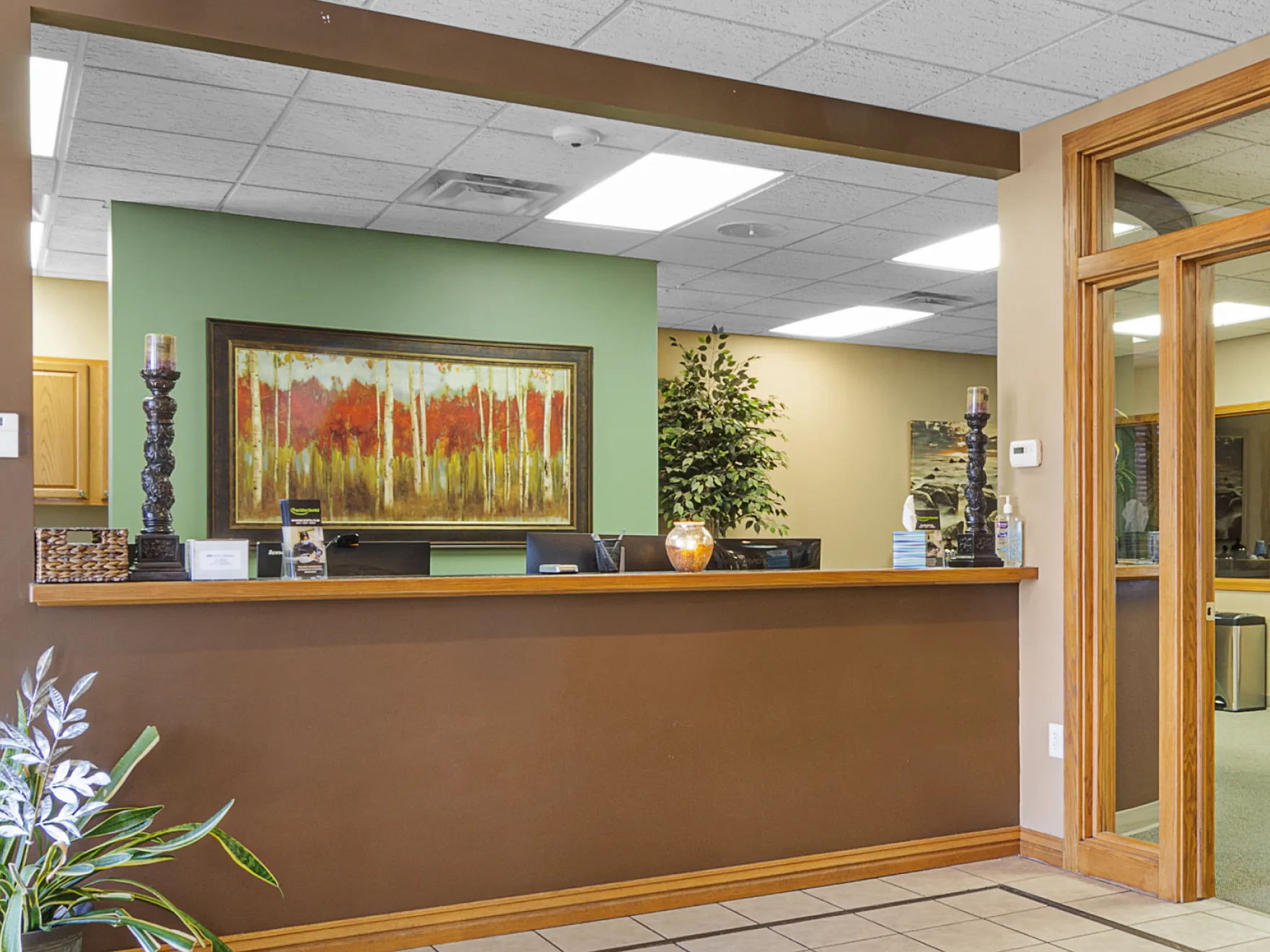 Reception desk area with green and brown walls, decorative candles, a large framed painting of red trees, potted plants, and office equipment behind the counter.