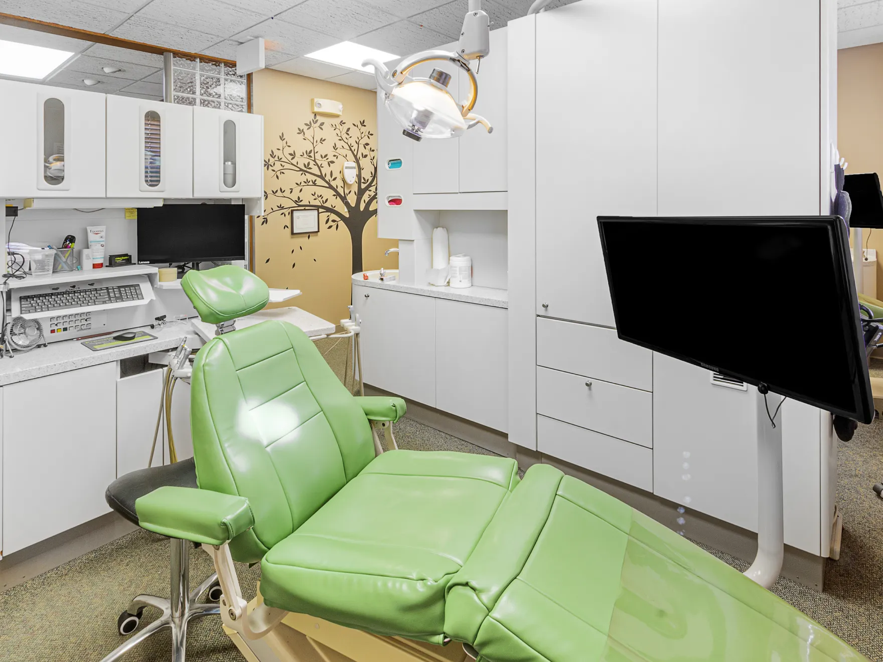 Dental treatment room with a green patient chair, mounted monitor, overhead dental light, and white cabinetry.