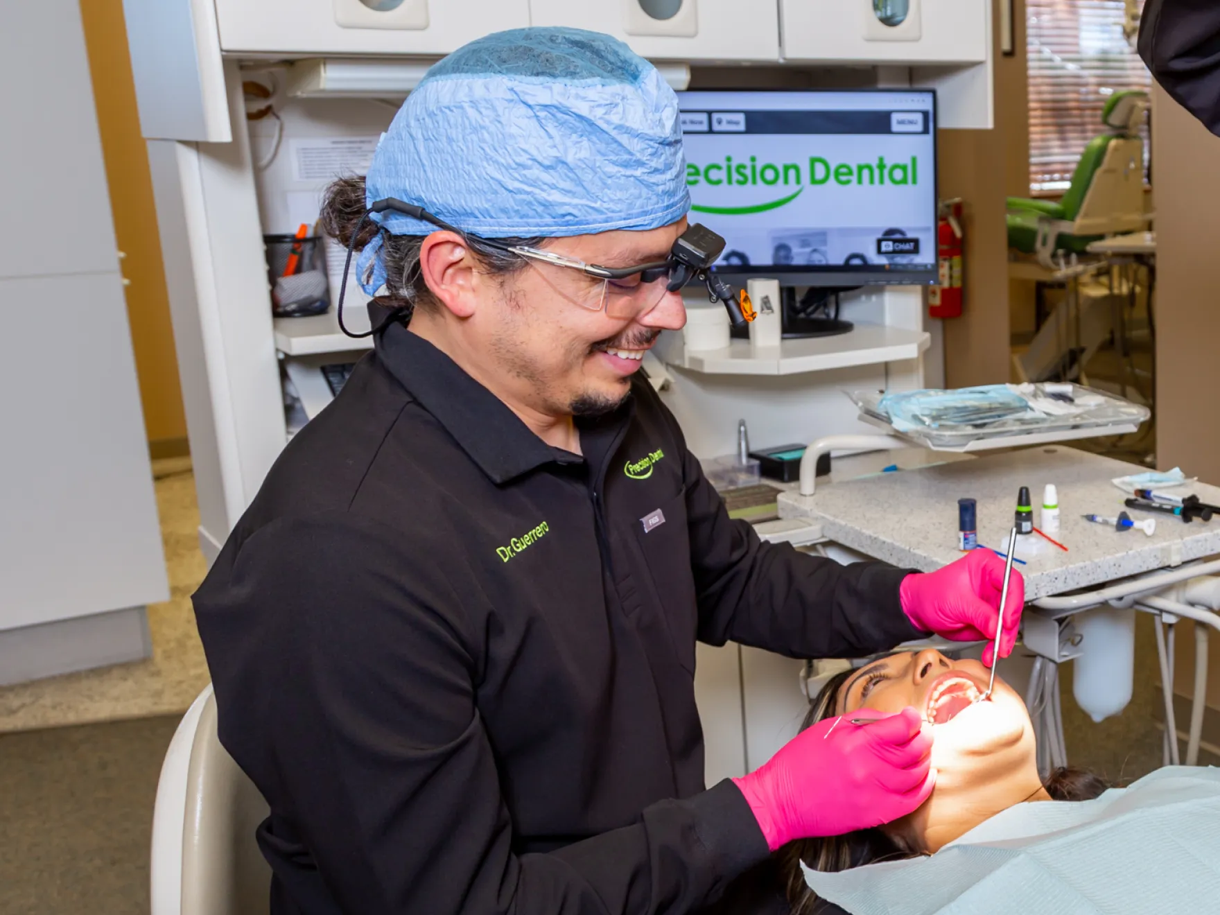 Dentist wearing pink gloves and a blue surgical cap examining a patient's teeth in a dental clinic.