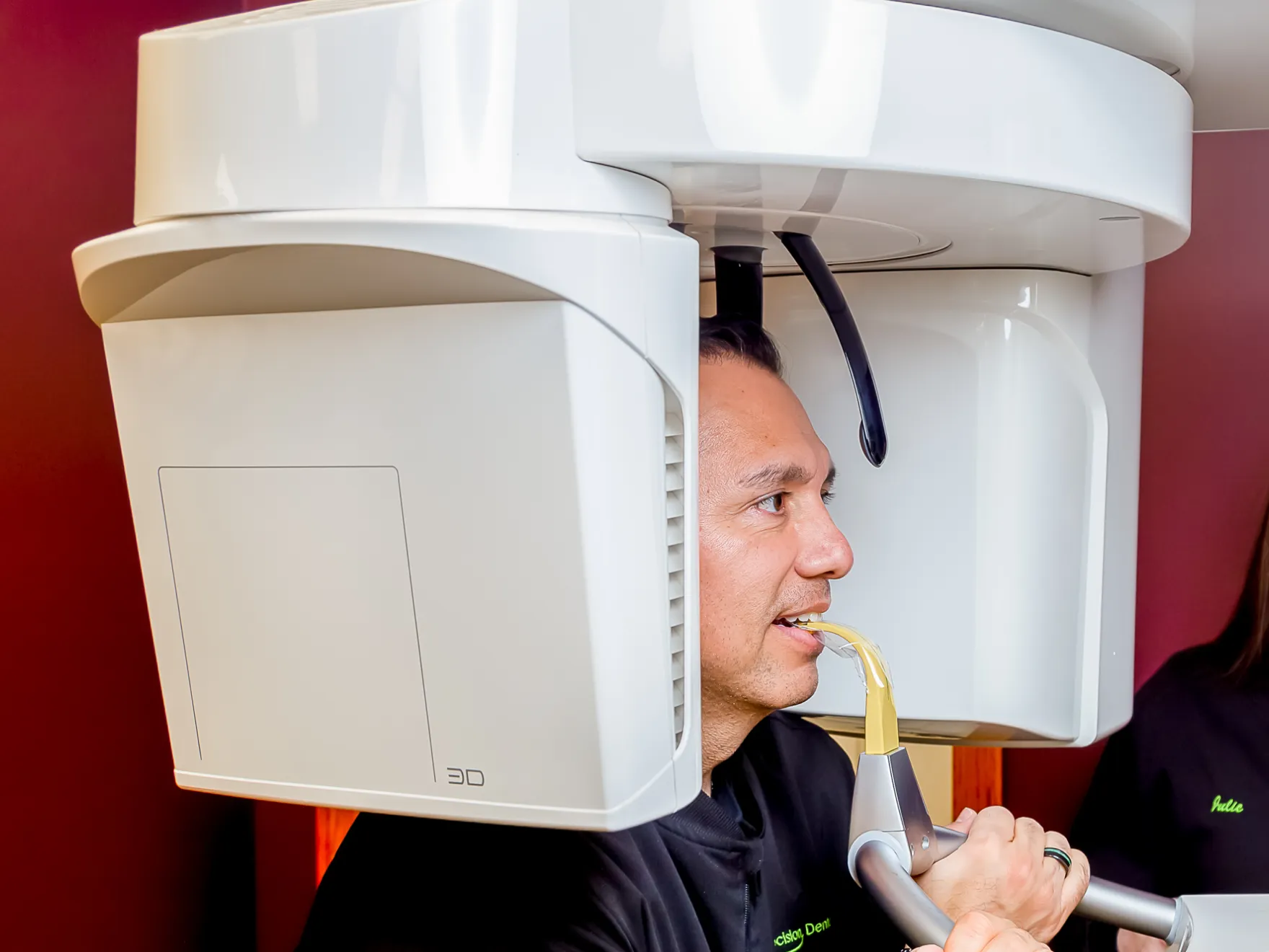 Man undergoing a 3D dental scan using a panoramic X-ray machine, biting on a yellow bite piece while holding onto the machine handles.