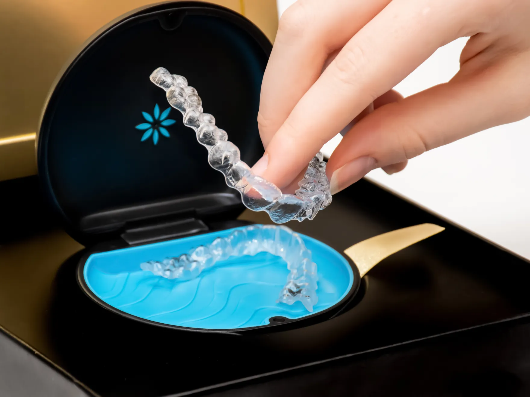 Hand holding a clear orthodontic aligner above a blue case inside a black and gold container.