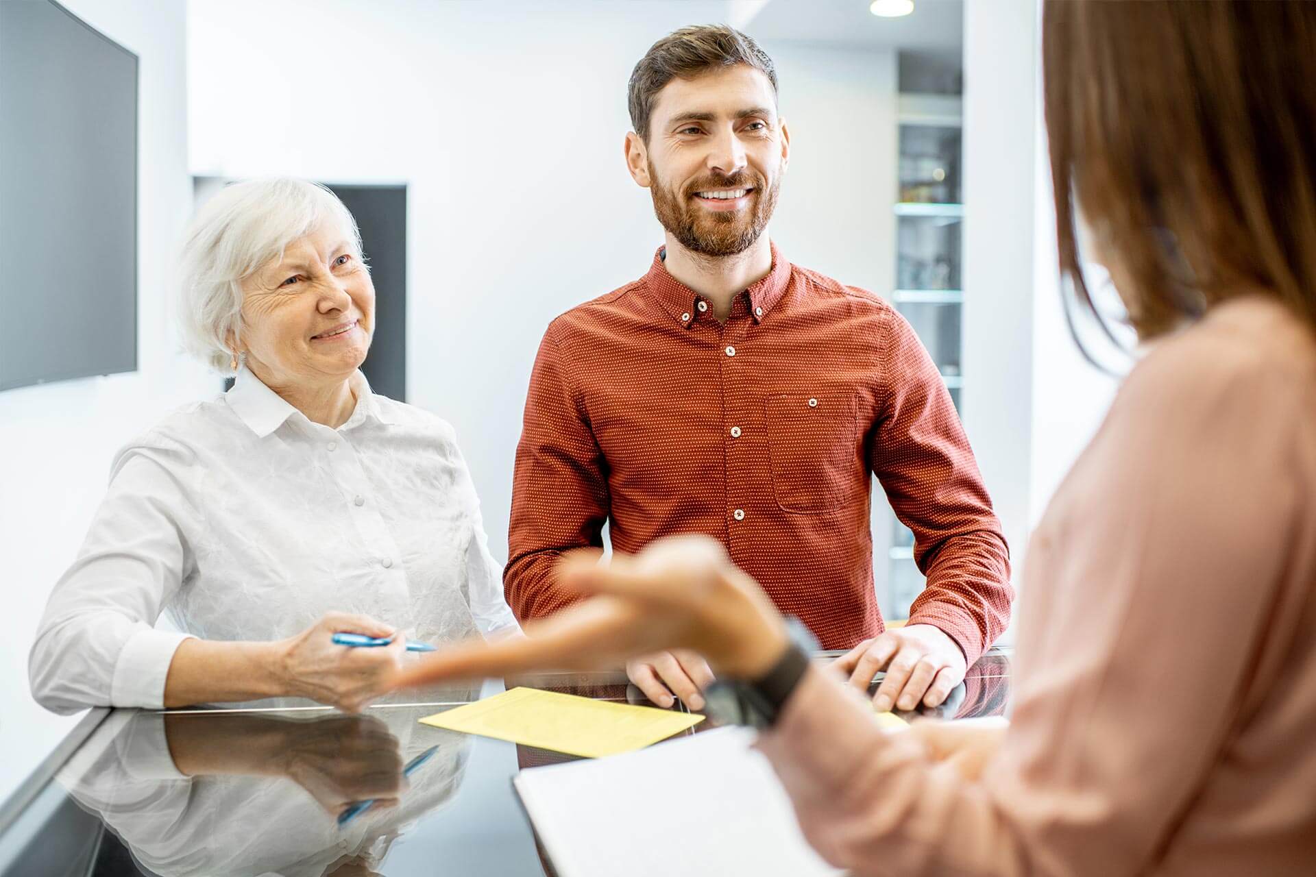 Smiling elderly woman and man in casual clothes having a discussion with a person holding a notepad across the table.